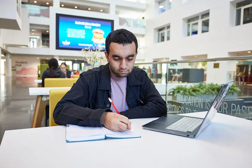 A student writing in a notebook with a laptop next to him.