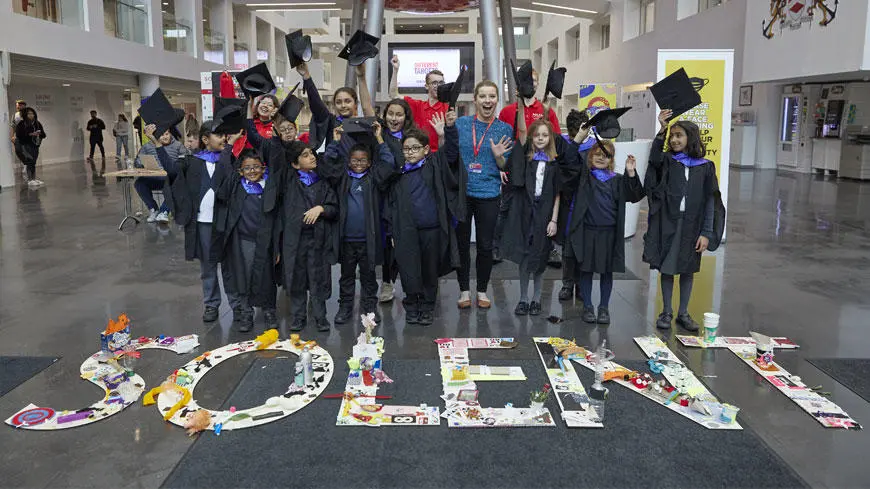 A group of school children wearing graduation caps and gowns behind the word SOLENT laid out on the floor