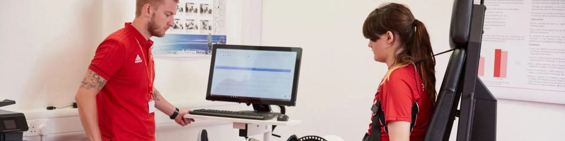 A female student sat down in a sport testing chair looks at a teacher beside a computer.