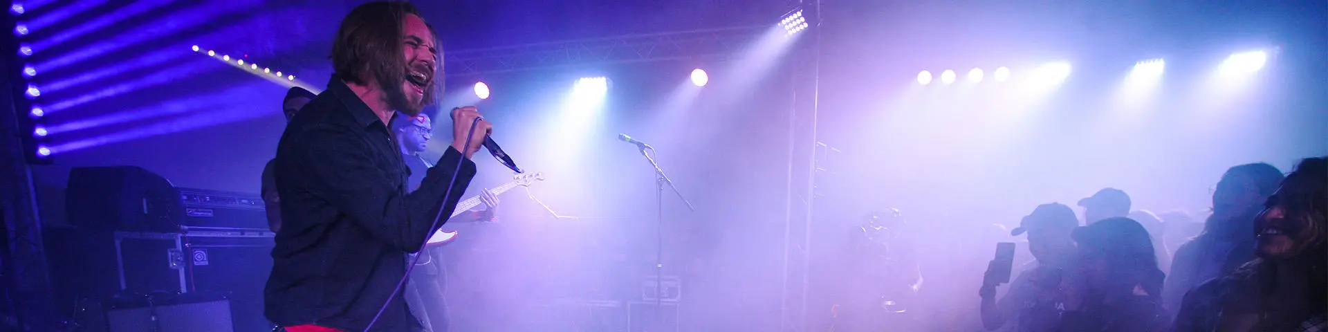 A male singer holds a microphone on a blue tinged stage whilst singing. In front of him is a crowd of people, some are holding mobile phones.