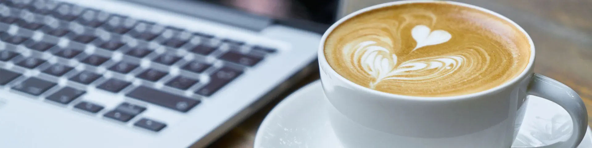 A full coffee cup with a heart in the top, next to a laptop on a desk.