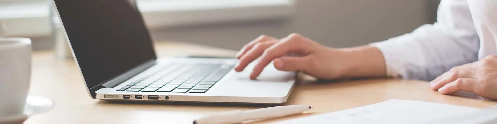 A hand rests on a light brown laptop on a desk, surrounded by a white mug, white pen and notepad.