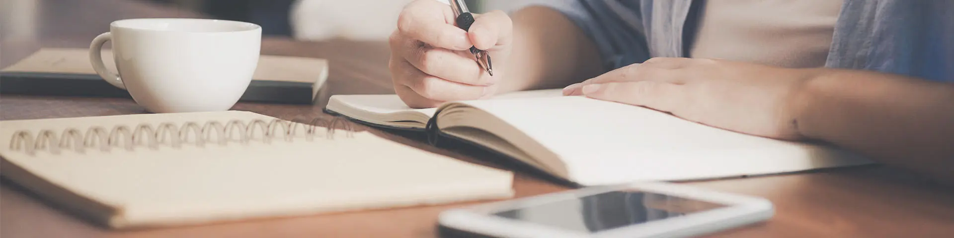 A hand writes on a notebook on a table surrounded by a cup and mobile